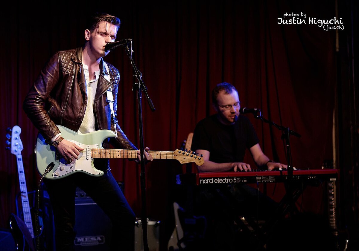 Patrick Droney playing a pale green electric guitar on a small stage, with a keyboardist behind him