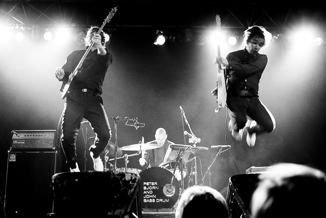 Black and white photo of two guitarists leaping on a smoky stage with a drummer behind them