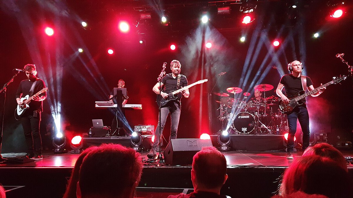 Five band members playing guitars, bass, keyboards and drums on a stage lit in red with beams of light, seen from behind the audience