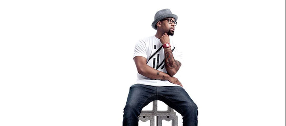 PJ Morton in a gray hat and glasses sitting on a stool against a white backdrop