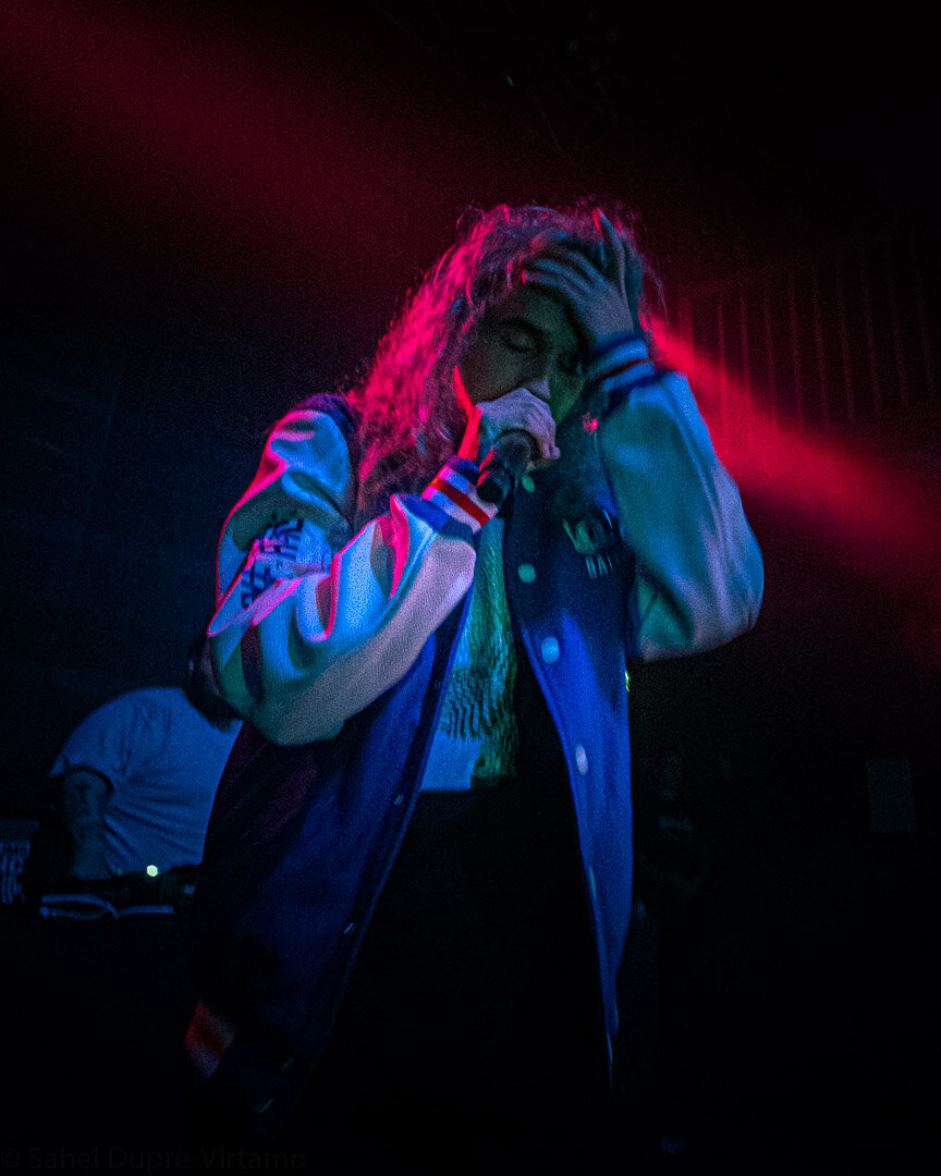 Rapper in a varsity jacket holding a microphone to his mouth under red and blue stage light