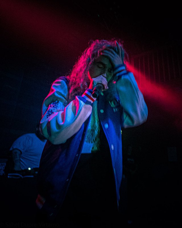 Rapper in a varsity jacket holding a microphone to his mouth under red and blue stage light