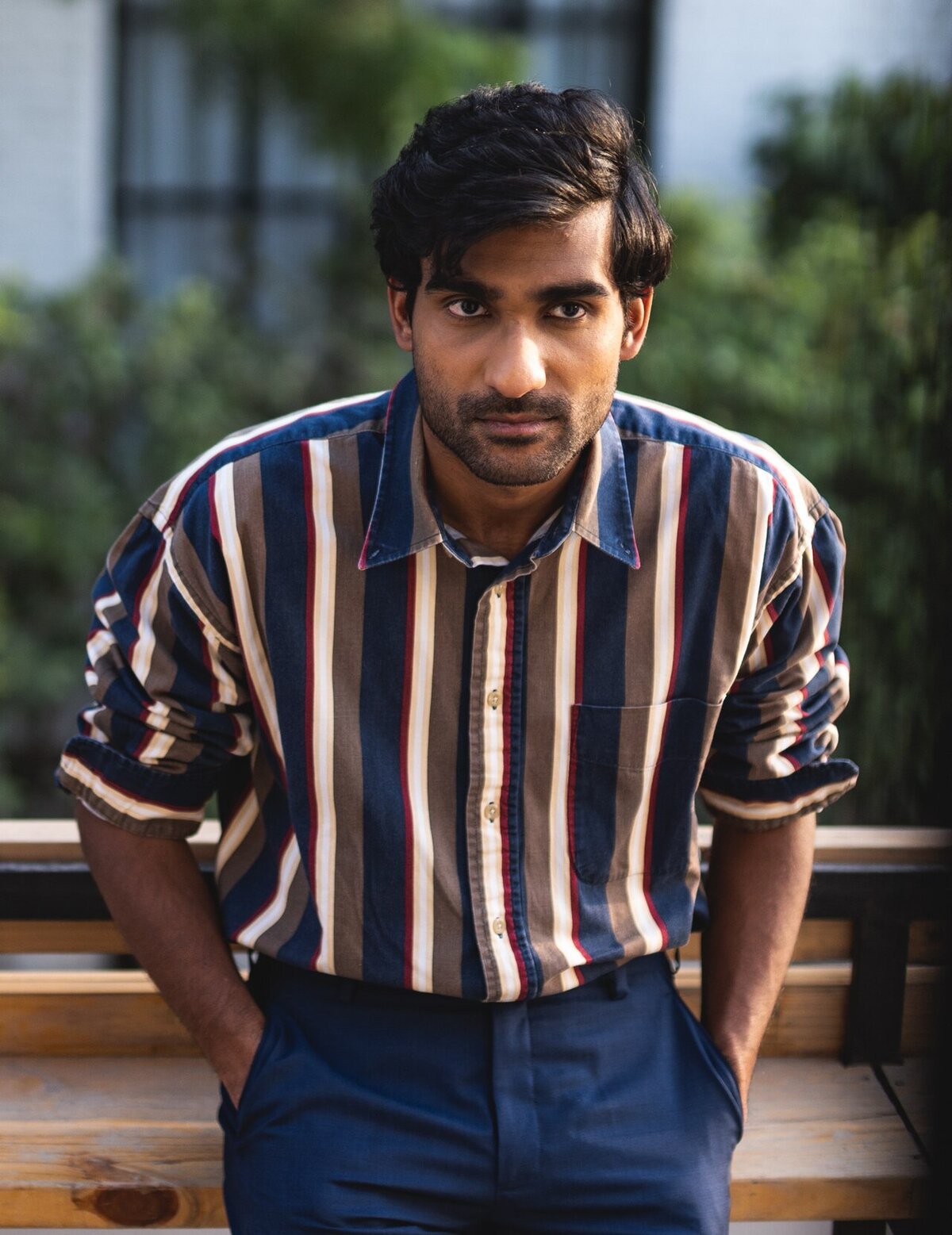 Portrait of Prateek Kuhad in a striped button-up shirt, hands in his pockets, standing in front of greenery