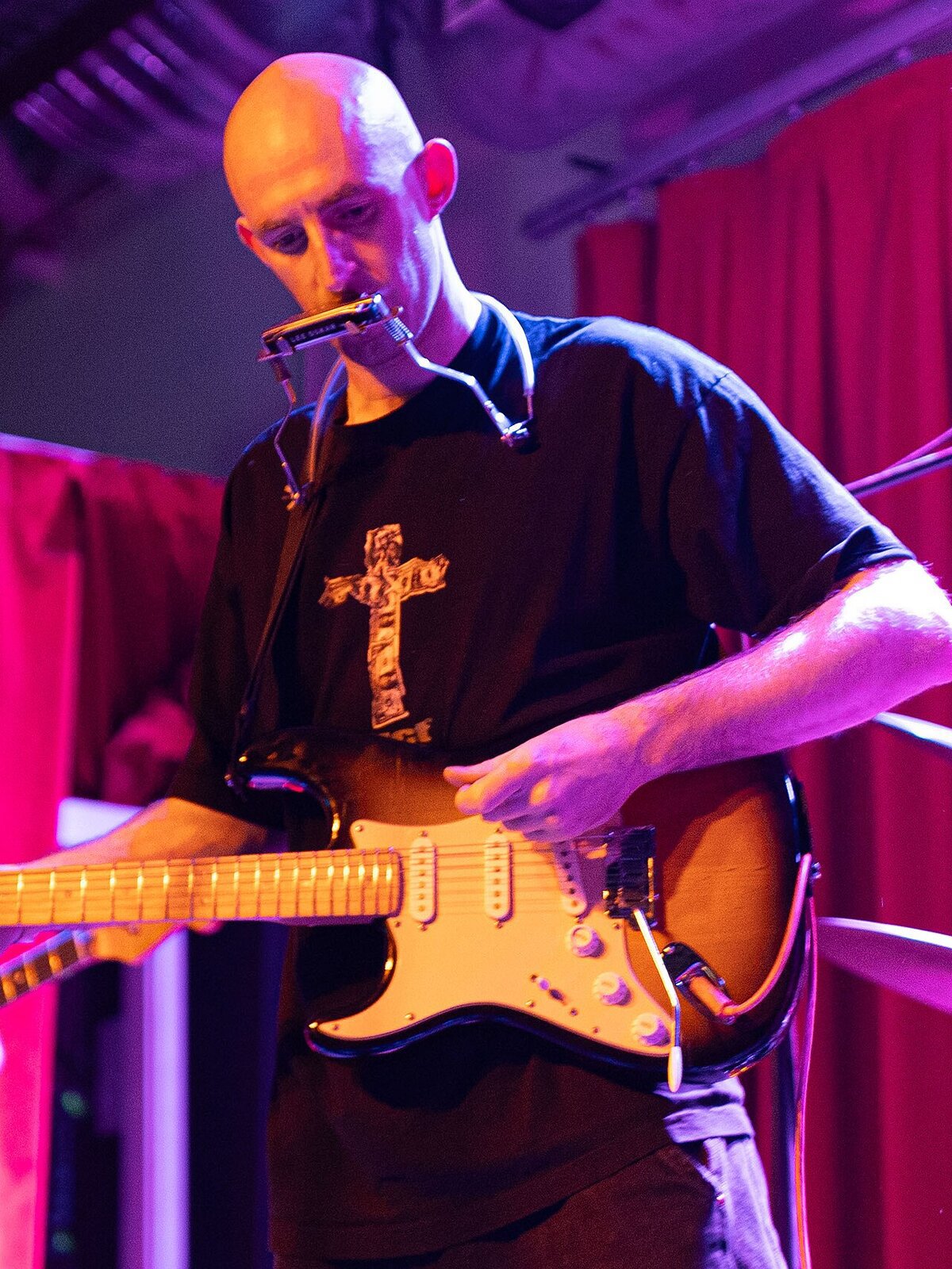 Musician with a harmonica rack playing an electric guitar under purple and red stage light