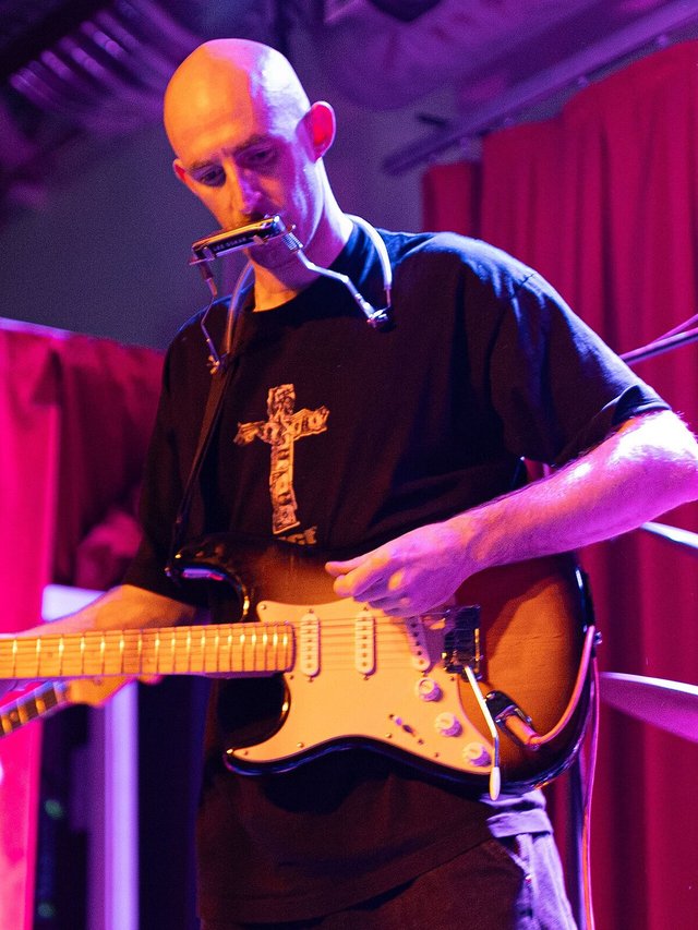 Musician with a harmonica rack playing an electric guitar under purple and red stage light