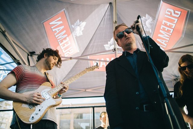 Protomartyr's singer in sunglasses at a microphone beside a guitarist during an outdoor daytime set
