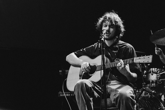 Black and white photo of Quadeca singing and playing an acoustic guitar at a microphone, with a drum kit behind him