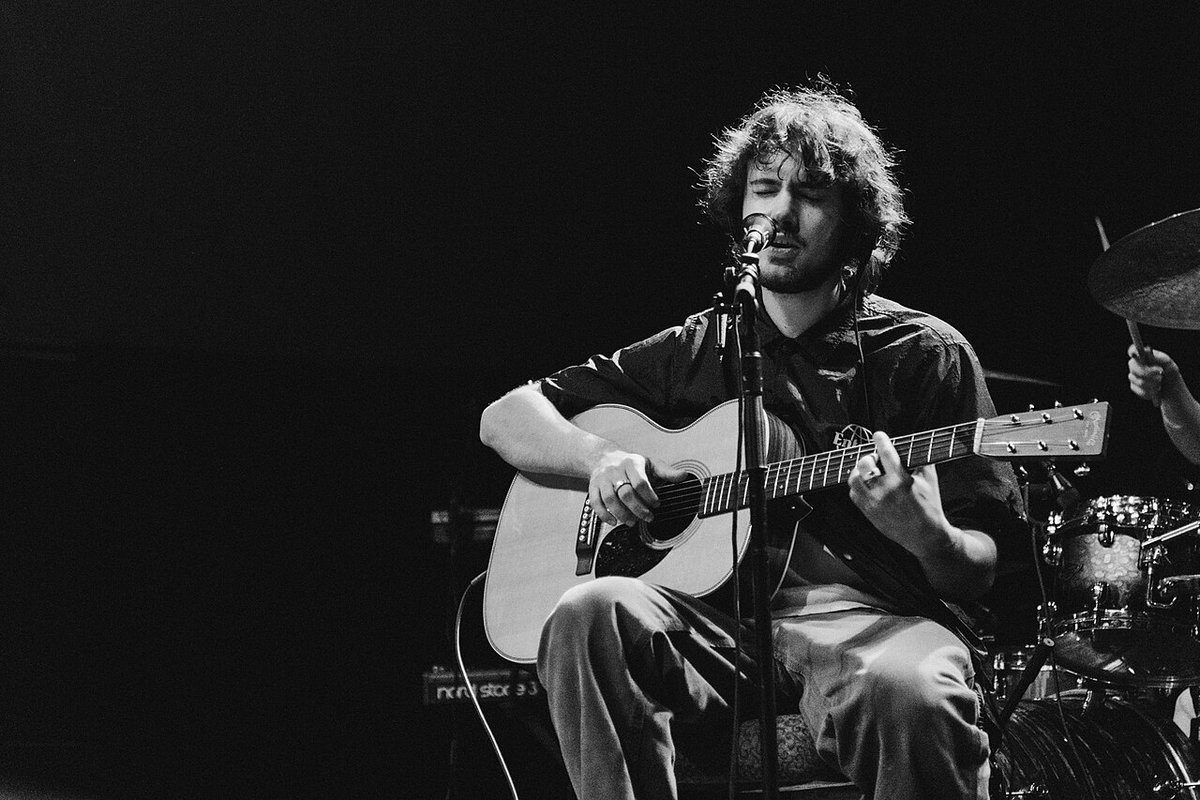 Black and white photo of Quadeca singing and playing an acoustic guitar at a microphone, with a drum kit behind him
