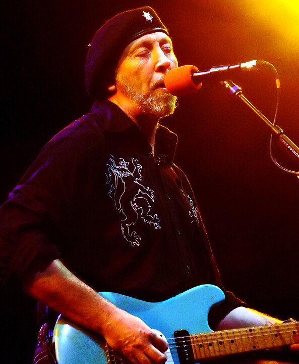 Richard Thompson singing into a microphone on stage while playing a light blue electric guitar