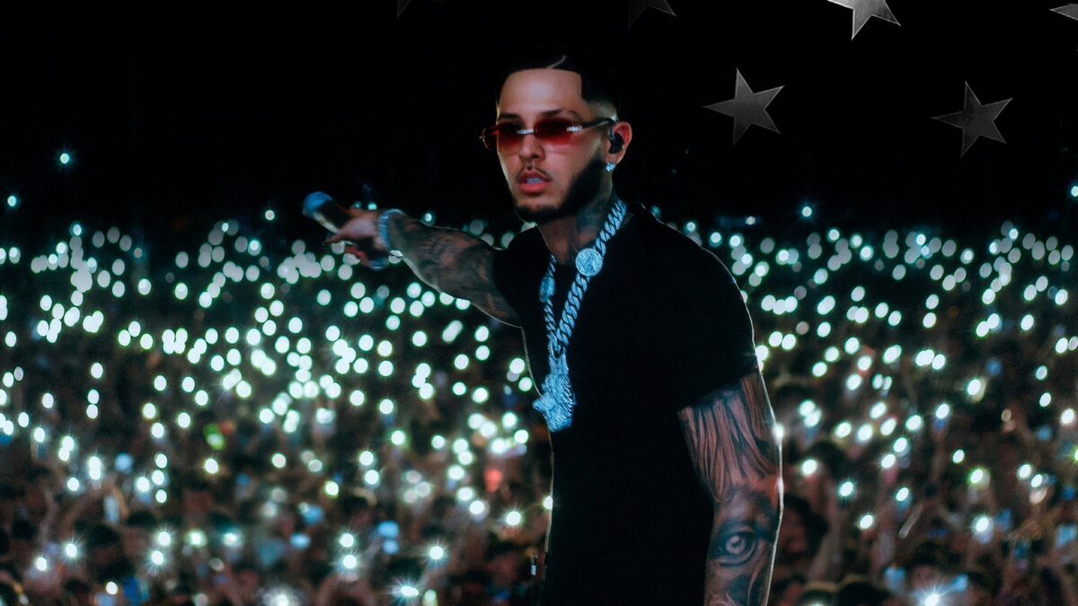 ROA in red-tinted sunglasses and a silver chain pointing a microphone toward a crowd lit by thousands of phone lights