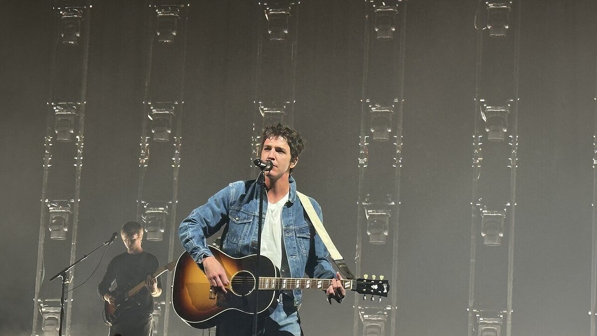 Singer in a denim jacket playing acoustic guitar at a microphone on a stage lit by tall columns of lights