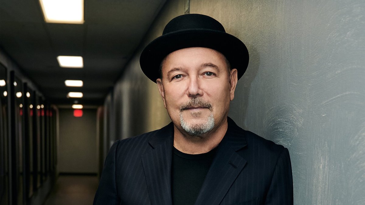 Rubén Blades in a black bowler hat and pinstripe jacket, leaning against a gray wall in a backstage hallway