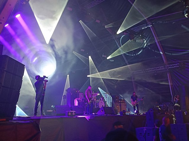 Three-piece band playing on a stage lit by purple and white beams of light, with a camera operator at left