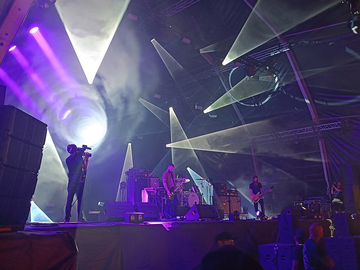 Three-piece band playing on a stage lit by purple and white beams of light, with a camera operator at left