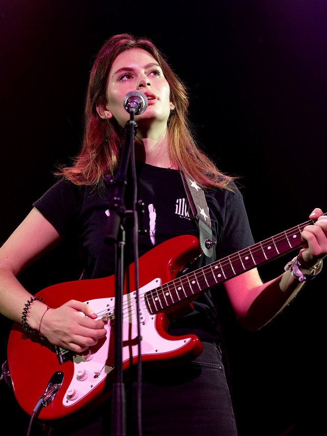 Singer in a dark jacket playing a red electric guitar at a microphone under red stage lights