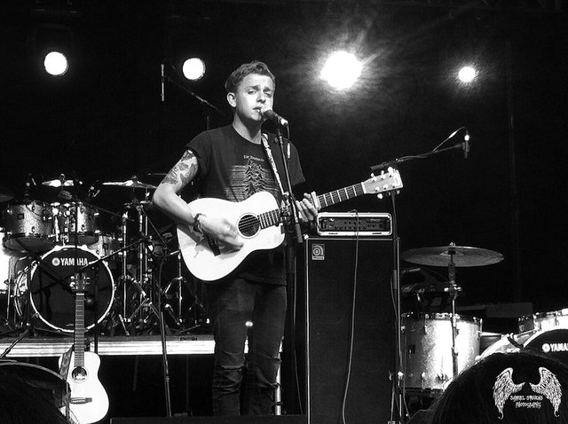 Black and white photo of a singer playing an acoustic guitar at a microphone on a dark stage with a drum kit behind him