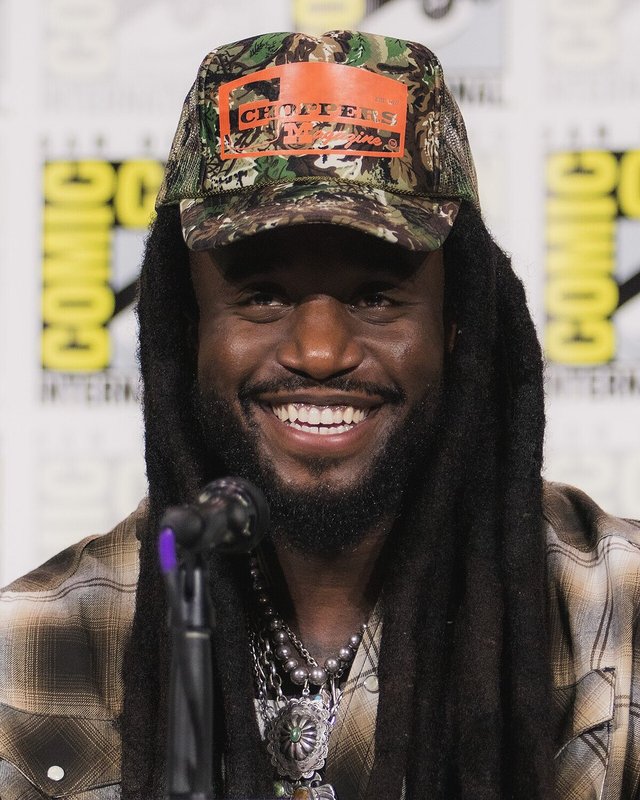 Smiling Shaboozey in a camouflage cap and plaid shirt seated at a microphone