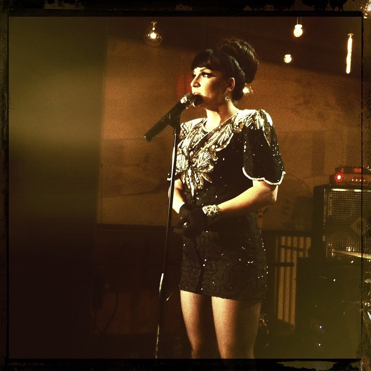 Singer in a sequined dress performing at a microphone stand on a warmly lit stage