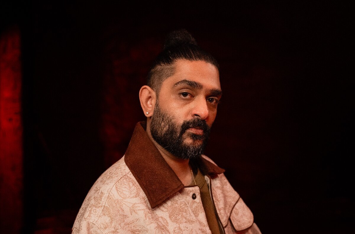 Portrait of a bearded man with a top knot in a pale patterned jacket with a brown collar against a dark red background