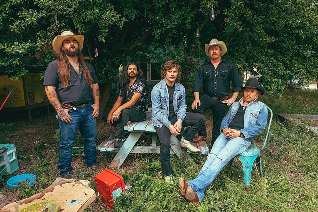Five band members in cowboy hats and denim posed around a picnic table under a tree