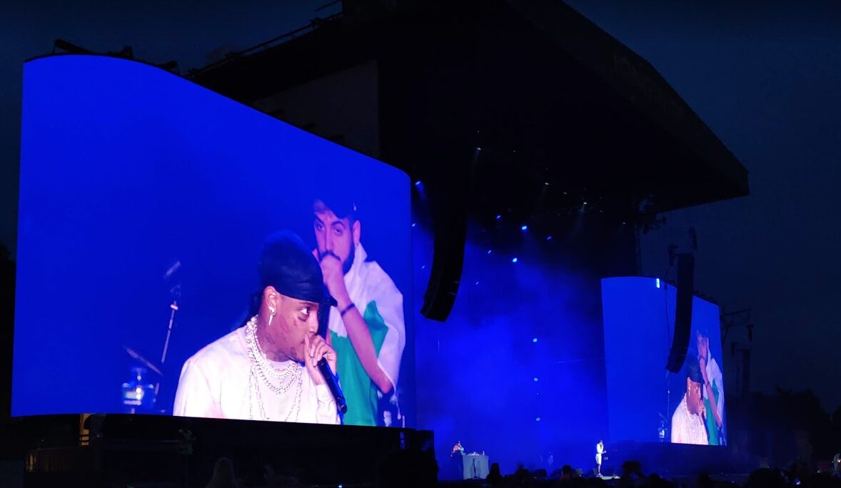 Rapper in a durag and chains on large blue video screens at a festival stage at night