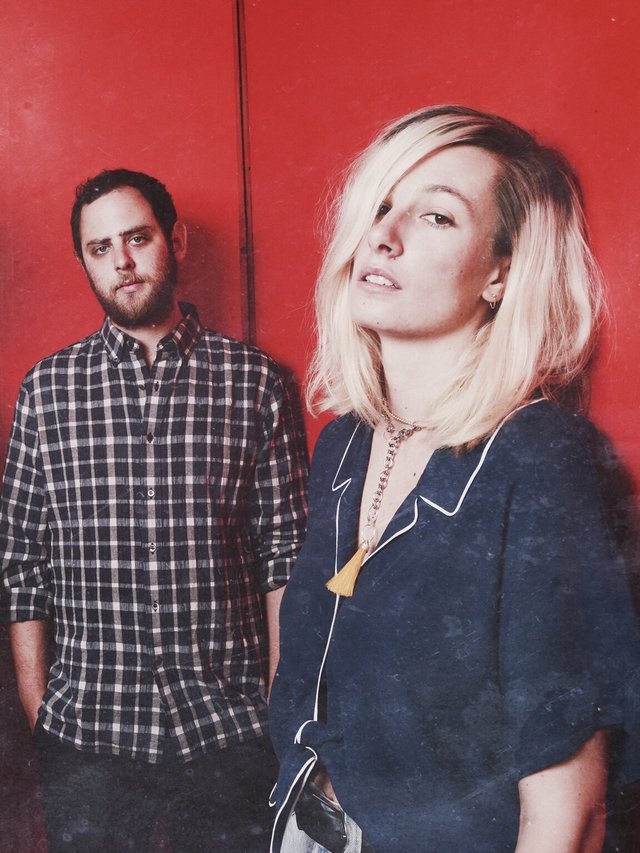 Two members of Slothrust posed against a red wall, a bearded man in a plaid shirt behind a blonde woman in a navy blouse