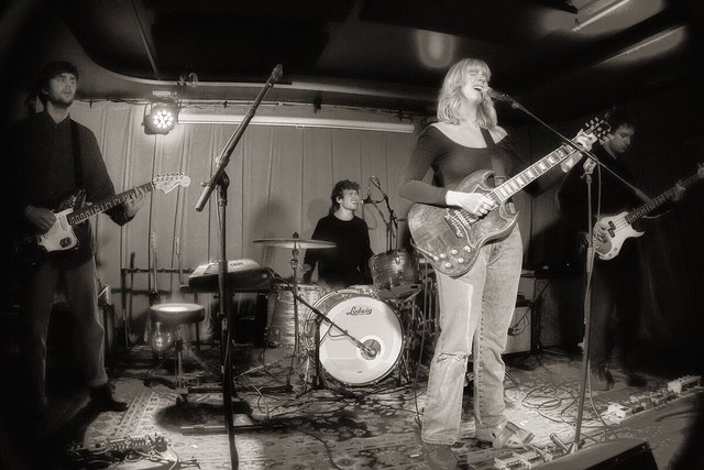 Black-and-white photo of a band on a small stage, a singer-guitarist in the foreground with a drummer behind her