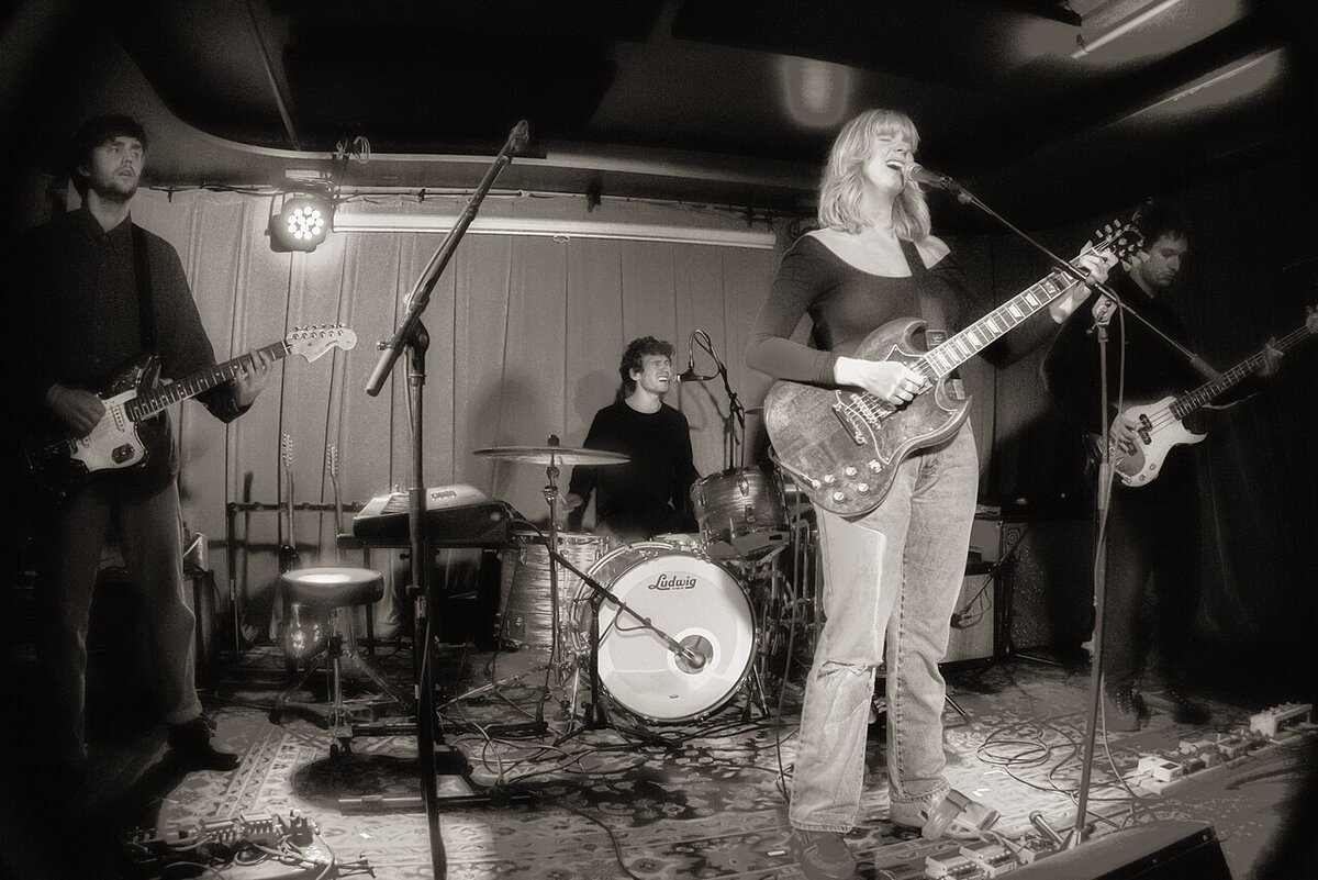 Black-and-white photo of a band on a small stage, a singer-guitarist in the foreground with a drummer behind her
