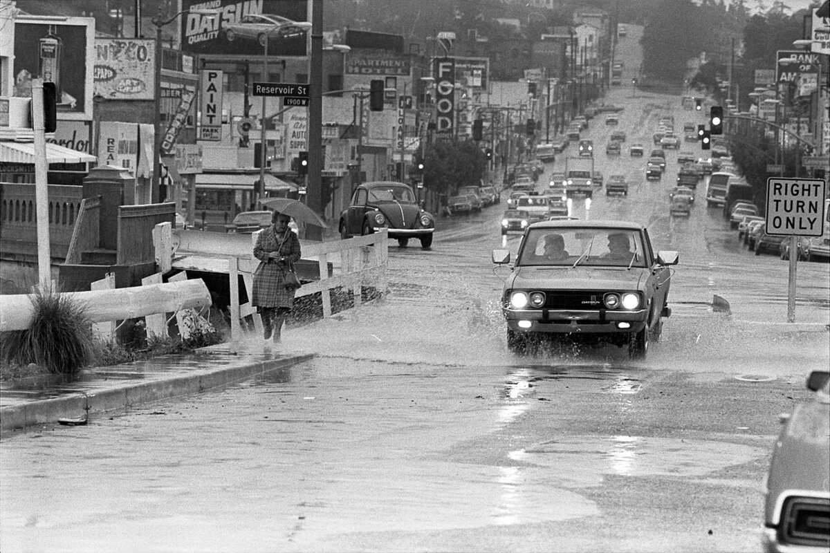Black-and-white street scene of a rainy Sunset Boulevard, a car splashing through a flooded intersection as a pedestrian with an umbrella walks past