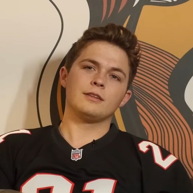 Josiah Queen seated against a mural wall during an interview, wearing a black football jersey
