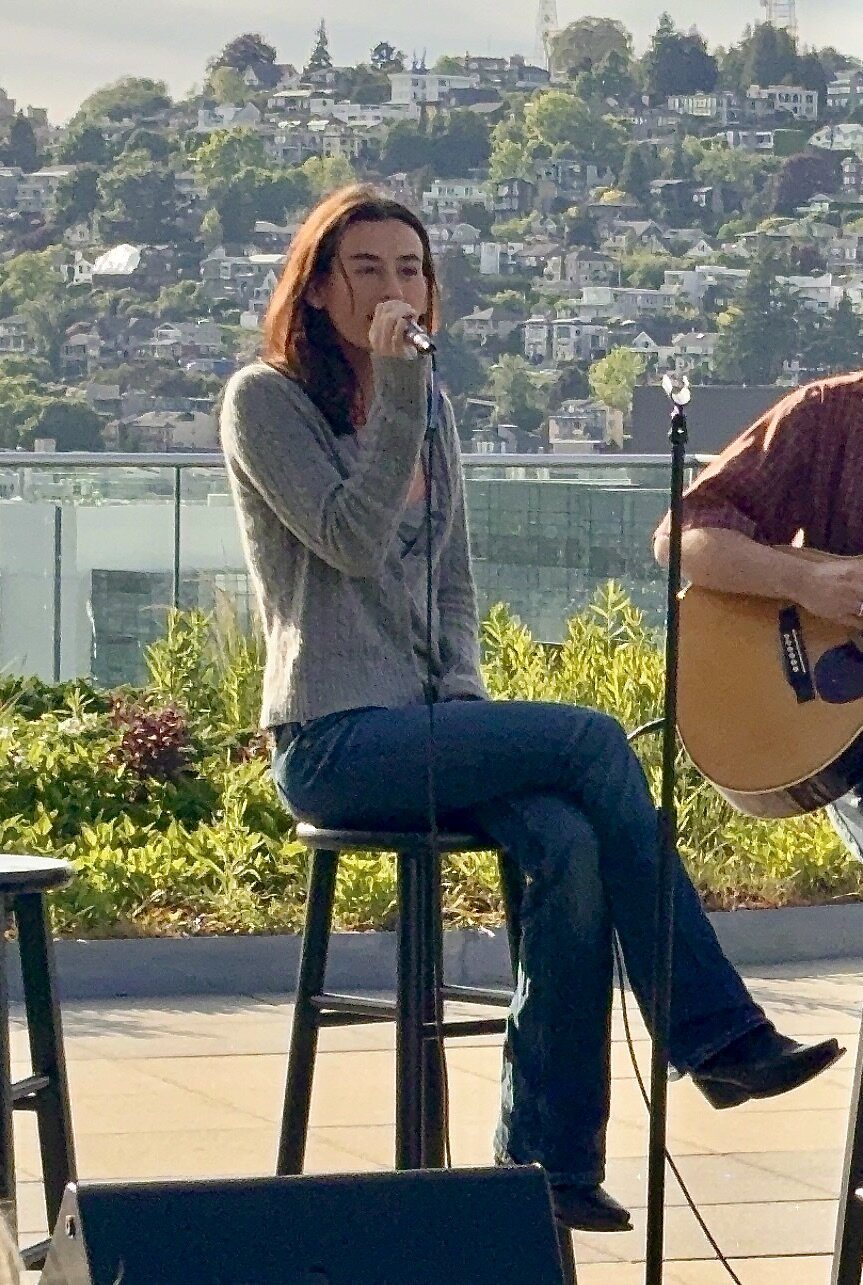 Singer seated on a stool singing into a microphone on a rooftop terrace, with a guitarist at the edge of the frame