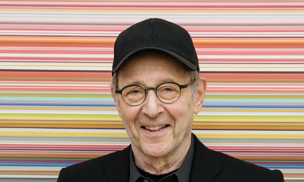 Smiling older man in a black cap and round glasses in front of a wall of colorful horizontal stripes