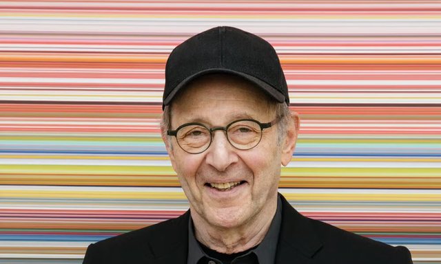 Smiling older man in a black cap and round glasses in front of a wall of colorful horizontal stripes