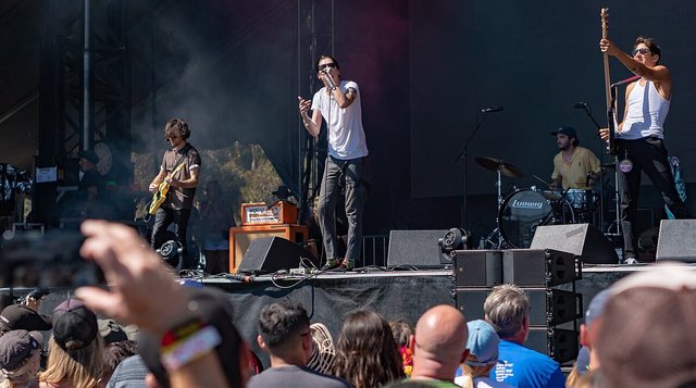 Plague Vendor performing on an outdoor festival stage, a singer at the microphone between a guitarist, a drummer and a bassist, with a crowd in the foreground