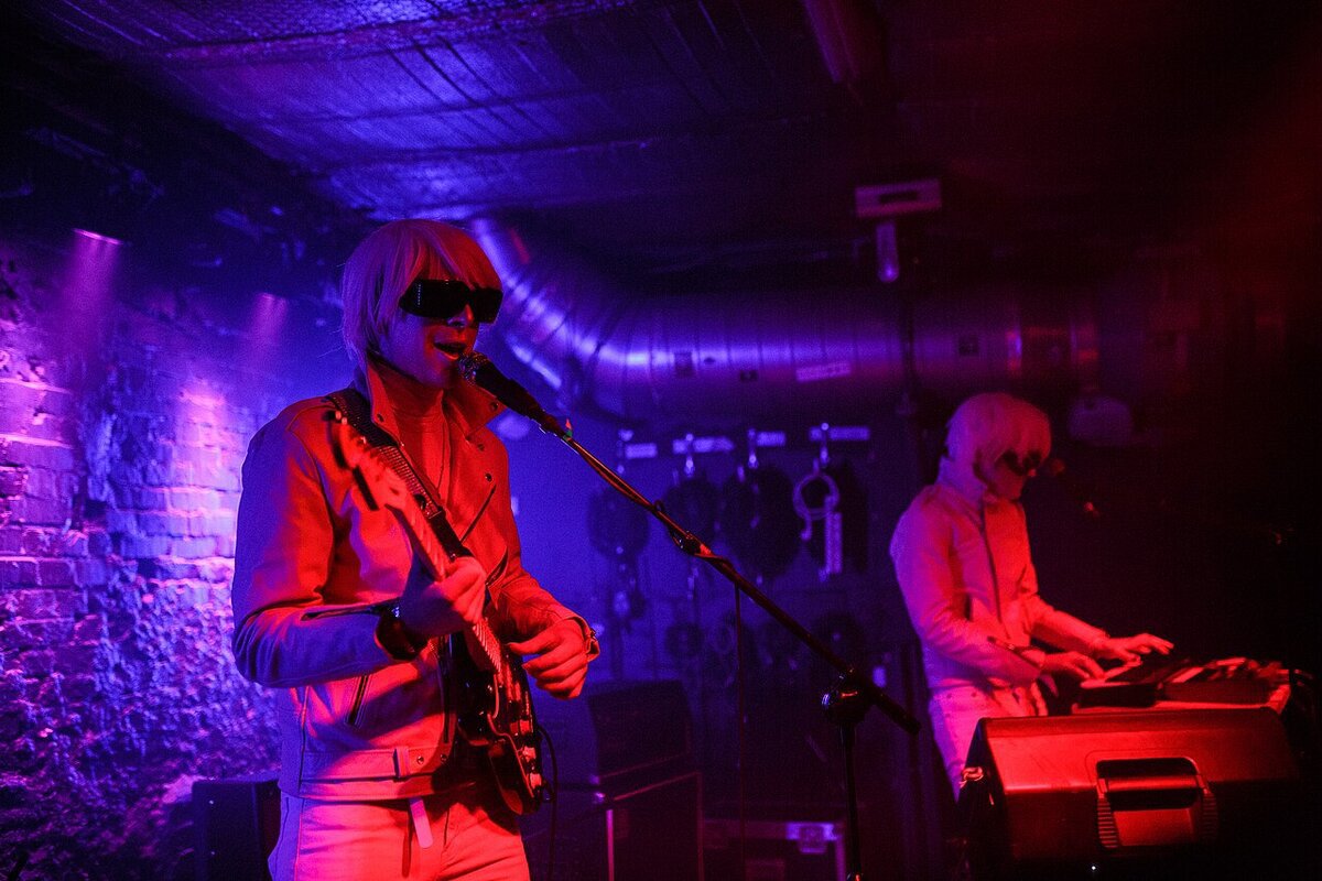Two performers in pale wigs and dark visors under red and purple stage light, one singing with a guitar, one at a keyboard