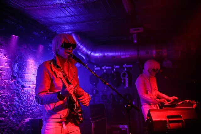 Two performers in pale wigs and dark visors under red and purple stage light, one singing with a guitar, one at a keyboard