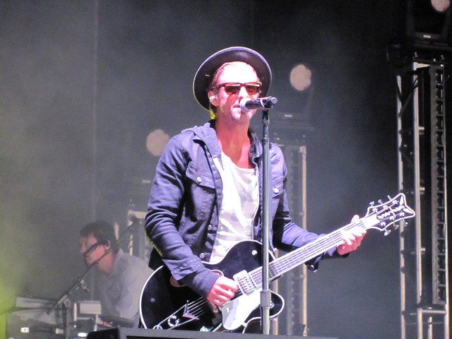 Switchfoot frontman Jon Foreman in a wide-brim hat and sunglasses, singing and playing a black electric guitar on a smoky stage