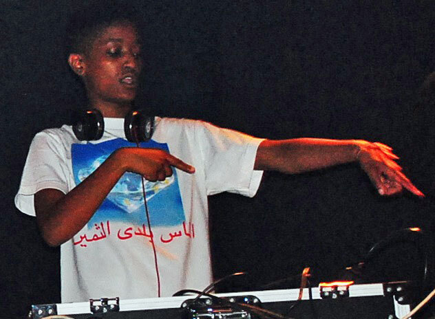 Syd standing behind a DJ table in headphones and a white graphic t-shirt, pointing off to the side
