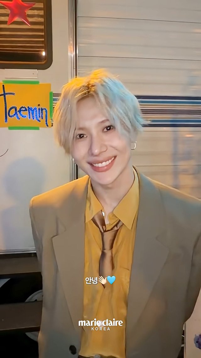 TAEMIN smiling in a tan blazer and gold shirt in front of a trailer door with his name on it