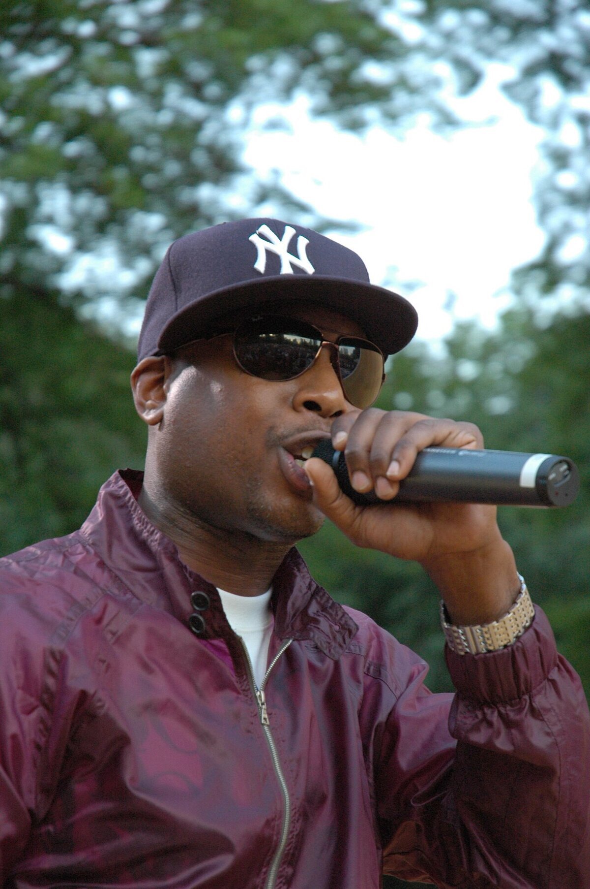 Rapper Talib Kweli performing outdoors in a navy cap, sunglasses and a maroon jacket, holding a microphone