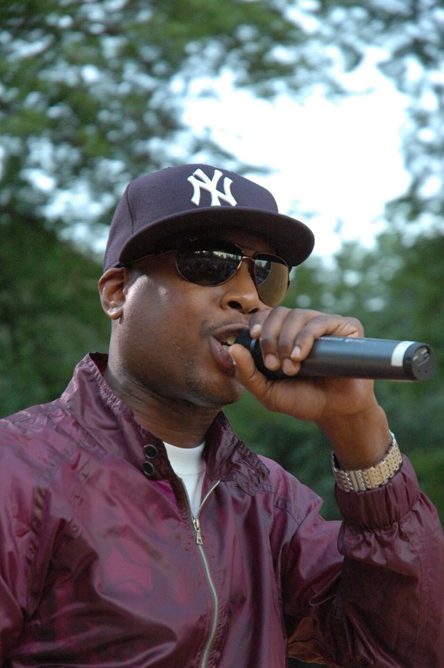Rapper Talib Kweli performing outdoors in a navy cap, sunglasses and a maroon jacket, holding a microphone