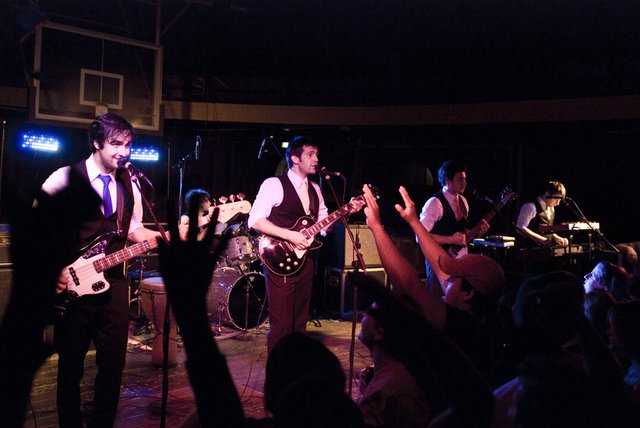 Band in vests and ties playing guitars and keyboards on a dim stage while fans raise their hands in the foreground