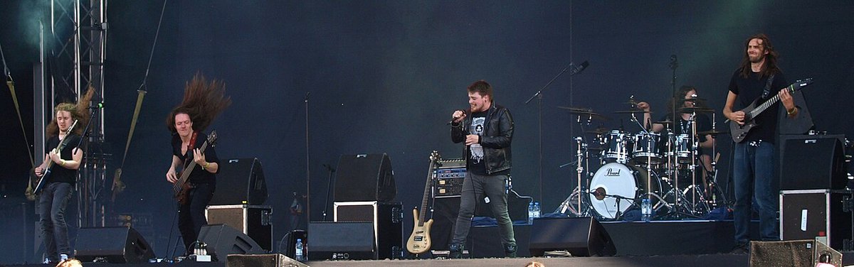 Five members of a metal band performing on a festival stage, one guitarist with long hair mid-swing and a singer at center