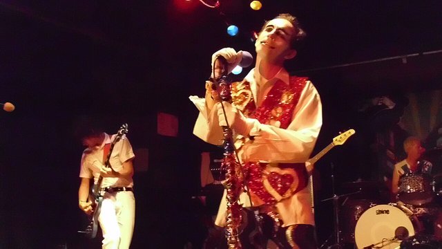 Singer in a sequined red vest and white gloves holding a microphone on stage, with a guitarist and drummer behind
