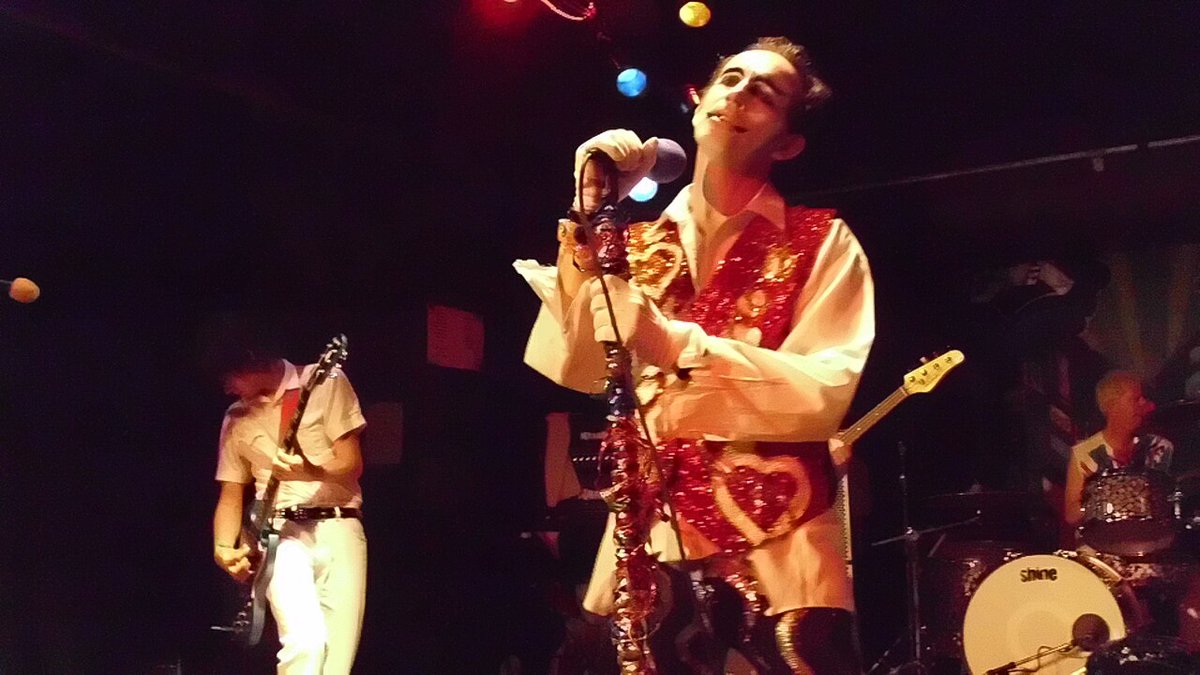 Singer in a sequined red vest and white gloves holding a microphone on stage, with a guitarist and drummer behind