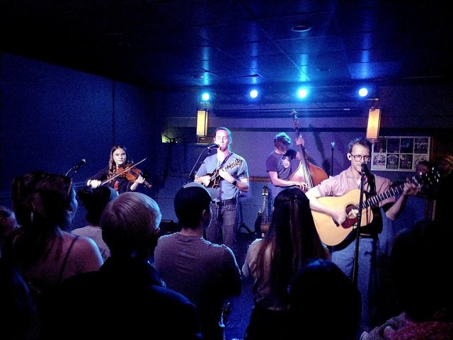 Four-piece folk band with fiddle, mandolin, upright bass and acoustic guitar playing to a small crowd under blue stage lights
