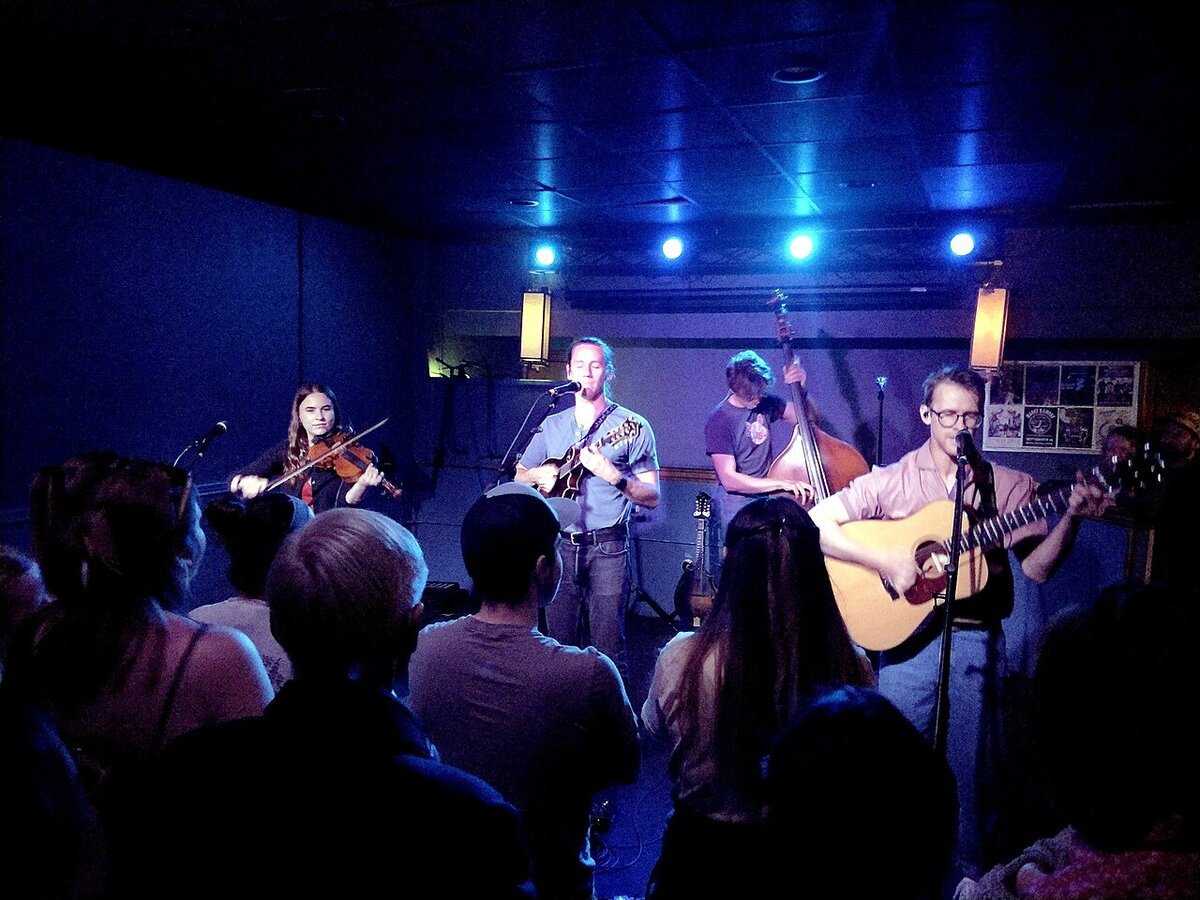 Four-piece folk band with fiddle, mandolin, upright bass and acoustic guitar playing to a small crowd under blue stage lights