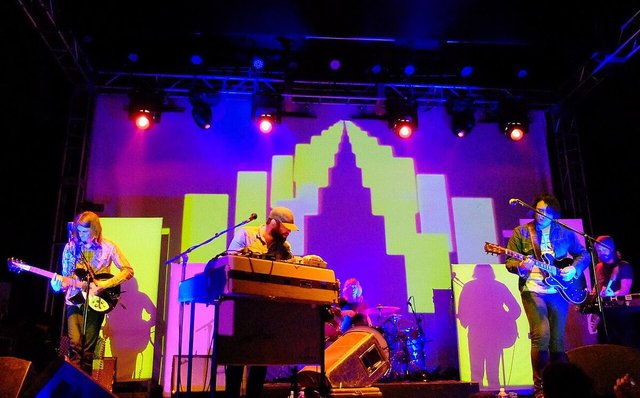 A band playing on a stage lit in purple and blue with a lime-green city skyline projected behind them