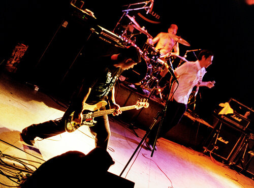 The Bouncing Souls playing on a dark stage under warm light, a bassist leaning forward and a singer at the mic