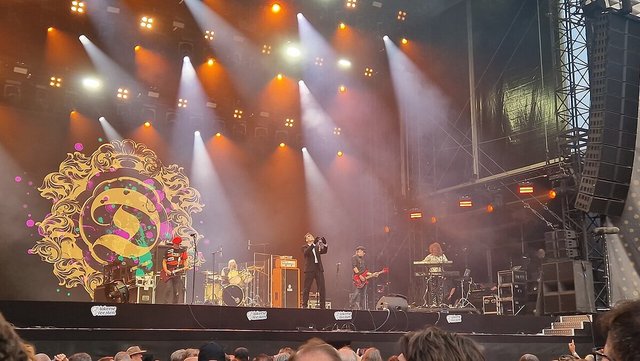 The Damned performing on a festival stage under orange and white spotlights, with the band's gold logo on a backdrop